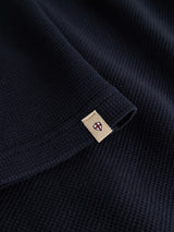 Close-up of the dark navy BGgreg L/S Grandad regular-fit cotton shirt by Blue de Gênes, showing its textured fabric and a beige rectangular tag with a small red and white emblem sewn onto the edge.