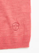 A close-up of the BGmonty Knit - Coral by Blue de Gênes, a crewneck jumper in a pink cotton-linen blend with a small shield and cross emblem stitched near the bottom hem, showcasing the fabric's texture and stitching details.