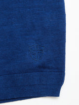 Close-up of the BGmonty Knit - Navy Peony by Blue de Gênes, showcasing its blue knit fabric with a small stitched shield emblem featuring a cross near the roll-rib hem. Crafted from a soft cotton-linen blend, the crewneck jumper is set against a white background.
