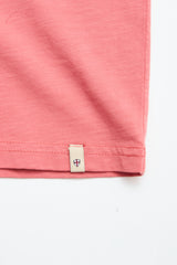 Close-up of the bottom hem of a coral BGoscar T-Shirt by Blue de Gênes in slub cotton, featuring a small beige tag with a dark shield emblem at the edge, set against a white background.