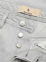 Close-up of BGgiacomo Color Jeans in Dolphin Grey by Blue de Gênes, made from rigid denim with silver buttons, a partially open fly, and a beige Blue de Gênes label featuring a small shield with a red cross.