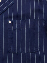 Close-up of the BGkendrick Toll Jacket - Navy Stripe by Blue de Gênes, a cotton worker jacket in navy blue with white vertical pinstripes. Featuring a chest pocket accented by a small, shield-shaped pin with a red cross on a white background near the pocket edge, this jacket offers a relaxed fit that combines comfort with timeless style.