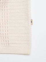 Close-up of the BGbilly S/S Cardigan in Chalk by Blue de Gênes—a cream-colored, 100% cotton piece with a textured knitted pattern and ribbed edges, featuring a small white tag with a purple cross symbol sewn into the seam.