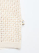 A close-up of the BGwalter S/S Cardigan in Chalk by Blue de Gênes showcases its cream-colored, 100% cotton waffle-knit fabric. A small beige tag with a red and blue shield emblem is visible on the seam, set against a plain white background.