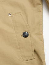 Close-up of the BGjacques Coat in Pale Khaki by Blue de Gênes, featuring a slanted pocket, black button, and a small shield-shaped pin above the pocket. The heavy cotton fabric appears smooth and sturdy, reflecting classic workwear inspiration.