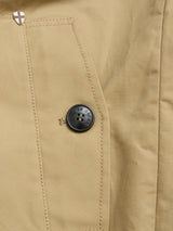 Close-up of the BGjacques Coat in Pale Khaki by Blue de Gênes, featuring a classic workwear pocket crafted from heavy cotton fabric. The pocket showcases visible stitching, a black button embossed with small text and symbols, and a small metallic shield-shaped detail at the upper left corner.
