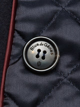 Close-up of a dark, glossy button featuring the Blue de Gênes logo, sewn onto quilted dark navy fabric from the Shiba Melton Coat. Diagonal stitch patterns and a maroon edge offer understated details that showcase Portuguese craftsmanship inspired by classic peacoat tailoring.
