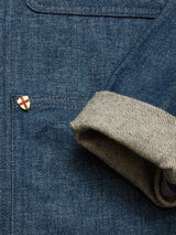 A close-up of the Blue de Gênes Yuma Fano Jacket in Denim Blue, featuring a relaxed fit, chest pocket with a small shield-shaped pin, and a rolled sleeve revealing striped inner fabric.