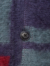 A close-up of the Block Knit Cardigan - Navy Pattern by Blue de Gênes shows its fuzzy, multi-color block knit fabric in purple, teal, and touches of red—reminiscent of a cozy wool blend—and features a four-hole black button with engraved text around the rim.