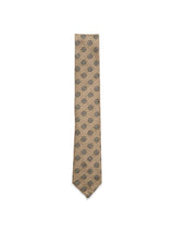 The Blue de Gênes Nekutaki Tie #E - Sand Pattern is a classic tan tie featuring a refined sand-patterned weave, with a repeating arrangement of circular black and gray floral-like motifs on a white background—an elegant men’s accessory for any wardrobe.