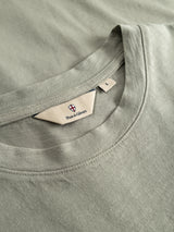 Close-up of the BGoscar T-Shirt in shadow green by Blue de Gênes, featuring a classic crewneck and a beige brand tag. Made from 100% cotton, the fabric appears soft with neat stitching.