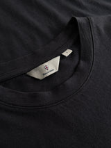 Close-up of a BGoscar T-Shirt - Jet Black by Blue de Gênes, featuring a white clothing tag with a small shield logo and text. The soft slub cotton fabric appears smooth and is neatly stitched around the crew neckline.
