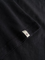 Close-up of a black crewneck fabric, likely the BGoscar T-Shirt - Jet Black by Blue de Gênes, made from 100% slub cotton, with a small white woven tag featuring a simple red and blue geometric design sewn into the hem.