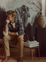 A man sits on a chair, resting his chin on his hand, wearing the BGenrico Hang Shirt - Navy Check by Blue de Gênes, crafted from 100% cotton with a classic collar, paired with tan pants. Beside him is a table with books, black skull decor, a large vase, and abstract black-and-white art in the background.
