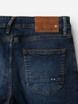 Close-up of the back of Blue de Gênes BGvinci Hermina Jeans - Med Used, crafted from dark blue comfort stretch denim. The regular fit jeans display orange stitching, a brown leather Blue de Gênes patch above the right pocket, and a small blue fabric tag on the pocket.