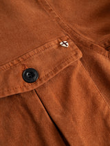 Close-up of the Blue de Gênes BGbarnaba Iro Overshirt in Ginger Bread Orange, a workwear-inspired garment crafted from soft comfort fabric for a relaxed yet durable feel. Details include a buttoned pocket and a small metal pin with a white background and black X.