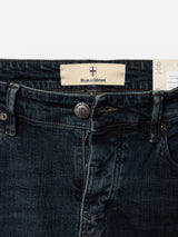 Close-up of the BGvinci Ewan Jeans - Color Denim by Blue de Gênes, regular fit in a dark blue wash with a visible button, zipper, and waistband label displaying the Blue de Gênes name and a small red and white shield logo above it. Made from comfort stretch denim to provide added ease of movement.