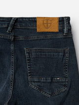 Close-up of the back of the BGvinci Ewan Jeans - Color Denim by Blue de Gênes in a dark blue wash, featuring a single back pocket and a tan leather patch with a subtly embossed shield logo near the waistband. Crafted from comfort stretch denim for ease of movement.