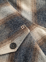 Close-up of the BGyodo Check Overshirt in Grey Check by Blue de Gênes, highlighting its plaid woolen fabric in beige, brown, and blue tones. The image features a metal button and a pin with a white shield and red cross design, showcasing the classic tailoring details of this men’s regular fit wool overshirt.