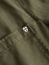 Close-up of the BGgabbiano Rory Blazer - Army by Blue de Gênes, crafted from washed herringbone fabric in olive green. The image highlights a pocket adorned with a small shield-shaped pin featuring a white background and a dark cross, positioned near the pocket seam.