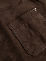 A close-up of the BGvalan Suede Jacket in dark brown by Blue de Gênes, highlighting a stitched pocket adorned with a small metallic pin featuring a white cross on a dark background near the top corner.