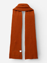 A BGcomforto Knit Scarf in Ginger Bread Orange by Blue de Gênes, featuring a long, ribbed chunky knit design with a small white tag and a rectangular patch near one end, is laid flat on a white background.