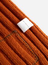 Close-up of the BGcomforto Knit Scarf in Ginger Bread Orange by Blue de Gênes, showcasing its textured orange wool fabric with a sewn white label reading 100% LAMBSWOOL, evoking the cozy warmth of a chunky knit accessory.
