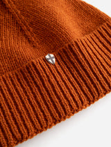 Close-up of the BGboshi Beanie in Ginger Bread Orange by Blue de Gênes, crafted from orange wool with a ribbed turn-up brim and adorned with a small silver shield-shaped pin featuring a black cross design—a tribute to timeless Blue de Gênes craftsmanship.