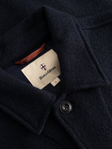 Close-up of the BGhew Wool Overshirt in dark navy by Blue de Gênes, size L, showcasing a visible button and a cream-colored label with the brand’s name and a small shield logo above it—evoking classic workwear style.
