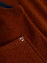 Close-up of the textured ginger bread orange fabric from the Blue de Gênes BGmudock Wool Waistcoat, with a small shield-shaped pin featuring a white background and black cross attached near the pocket’s edge.