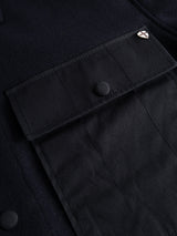Close-up of the BGBoyd Wool Jacket in Dark Navy by Blue de Gênes, showing a utility pocket with a flap and button closure. A small metal pin with a shield design is attached near the top corner of the pocket, emphasizing the jacket’s durable workwear aesthetic and understated waxed cotton accents.