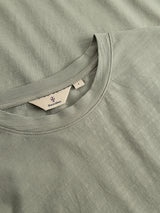 A close-up of a shadow green BGoscar T-Shirt highlights the crewneck collar and a tag featuring the Blue de Gênes brand and size L, displayed on a flat, matching slub cotton fabric background.