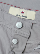Close-up of flint gray BGpaulo Teo Pant by Blue de Gênes, featuring metallic buttons and a label marked "Blue de Gênes" above a red and white shield logo on a beige background. The visible stitching around the waistband and button area evokes the look of vintage military pants, emphasizing the durability of their chino construction.