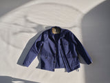 The BGcanopy Jacket in Navy Peony by Blue de Gênes is laid flat on a light surface, its rich blue shade, buttoned front, and multiple pockets highlighted by soft shadows and sunlight that cast a gentle pattern, evoking classic workwear heritage.