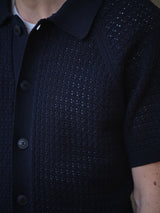 A close-up of a person wearing the BGbilly S/S Cardigan in Dark Navy by Blue de Gênes—a short-sleeve, textured knit button-up shirt with a collar. Crafted from 100% cotton and featuring vintage-inspired detailing, only part of the chest, arm, and neck are visible.