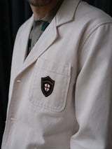 A person wearing the Blue de Gênes BGbaio Twill Blazer in Chalk, featuring a chest pocket patch with a shield, cross, and “Union des Créatifs” text. The background is dark and the person’s face is partially out of frame.