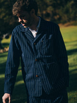 A man stands outdoors on grass in sunlight wearing the Blue de Gênes BGkendrick Toll Jacket - Navy Stripe, a relaxed fit cotton jacket featuring blue and white stripes. He looks down with his left arm lowered as partial shadow falls across his face, with blurred trees in the background.