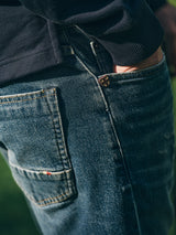A close-up of a person wearing Blue de Gênes BGvinci Striker Jeans - Dark Distress, crafted from comfort stretch denim in a regular fit, styled with a dark jacket and one hand in their pocket. The jeans feature a small embroidered patch near the pocket.