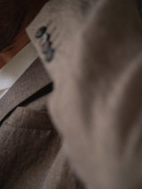 Close-up of a person wearing the BGcricket Lin Blazer in Grey Mel by Blue de Gênes, styled with a white collared shirt. The relaxed fit blazer features a visible suit lapel and subtle button details, captured in soft lighting with part of the neck and jawline showing.