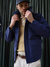 Clad in the Blue de Gênes BGcanopy Jacket in Navy Peony, along with a beige and yellow striped sweater, white pants, and a navy cap, a person stands before dark green curtains, adjusting the collar of the jacket.