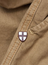 A close-up of Blue de Gênes BGpaulo Pavia 1 V2 Chino - Fungi, brown cotton stretch fabric with orange stitching and a small shield-shaped pin with a white background and red cross, inspired by vintage military pants.