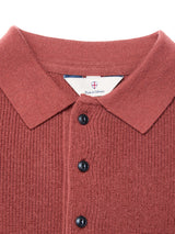 Close-up of the Porto Polo Knit in Henna by Blue de Gênes—a rust-colored, ribbed knit polo with a collar and black buttons. A small tag inside the collar features the shield logo and brand name.