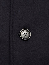 A close-up of the Shiba Melton Coat in dark navy by Blue de Gênes showcases a round, four-hole button inscribed with the brand name and featuring a small shield symbol beneath the text. The coat’s fabric appears to be high-quality wool or similar material, reflecting the exceptional craftsmanship associated with Portuguese tailoring.