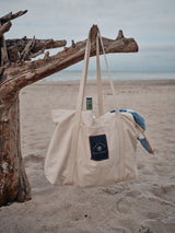 The BDG Canvas Bag - Off White by Blue de Gênes hangs from driftwood on a sandy beach, holding a green bottle and a blue-and-white towel, with the sea and cloudy sky in the background.
