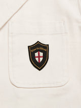 A close-up of a pocket on the BGbaio Twill Blazer in Chalk by Blue de Gênes, made from white cotton twill fabric and detailed with a black and gold shield-shaped patch featuring a red cross and the Blue de Gênes logo.