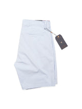 The Menzino Shorts - Blue Water by Blue de Gênes are light blue cotton-linen chino shorts, neatly folded with a dark tag on a string, displayed against a white background.