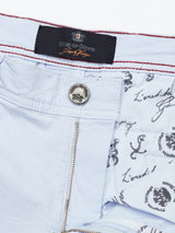 Close-up of Blue de Gênes Menzino Shorts - Blue Water: light blue cotton-linen blend, metal button, zipper, inner lining with script and crest patterns, and a black label with red stitching sewn at the waistband.