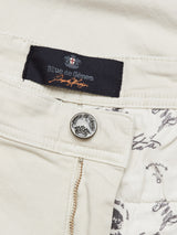 Close-up of Menzo Dobby Chino - Stone by Blue de Gênes, featuring a crown-embossed metal button, zipper, branded label, and fabric lining with black script and vintage-inspired patterns.