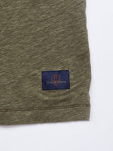 A close-up of the hem of the Totti T-Shirt - Army by Blue de Gênes, showcasing its olive green textured fabric and a small navy patch with an orange-outlined emblem and "mad genes" stitched on it.