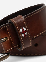 A close-up of the BGualan Belt - Brown by Blue de Gênes, crafted from full-grain leather and handmade in Italy, featuring distinctive white stitching and a small shield emblem with a red cross on a white background. The classic metal buckle lends a timeless touch to this finely crafted accessory.